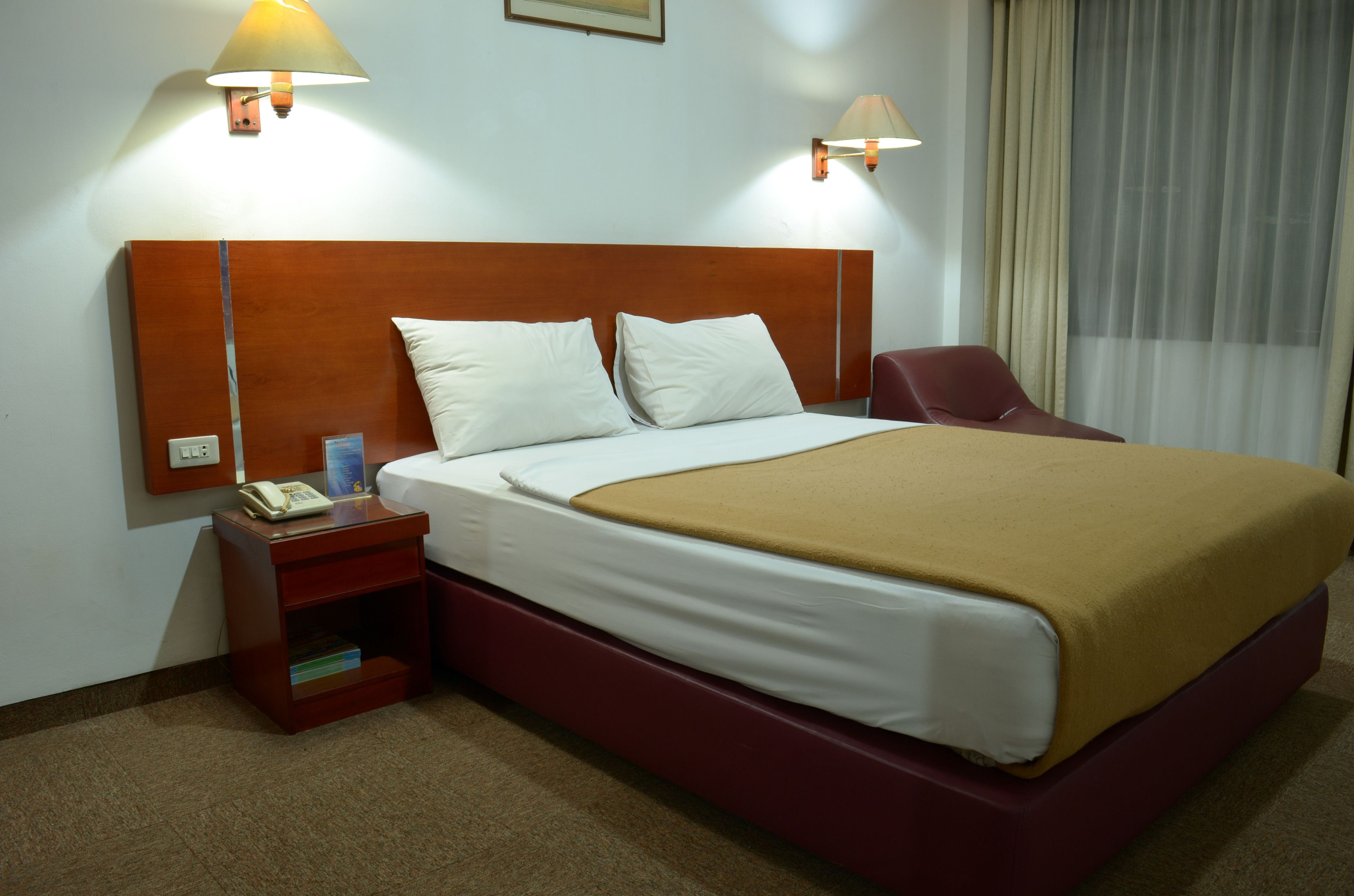 Une chambre d’hôtel avec une tête de lit en bois, deux oreillers blancs, un plaid marron, une chaise rouge, une table de chevet avec une lampe et un téléphone.