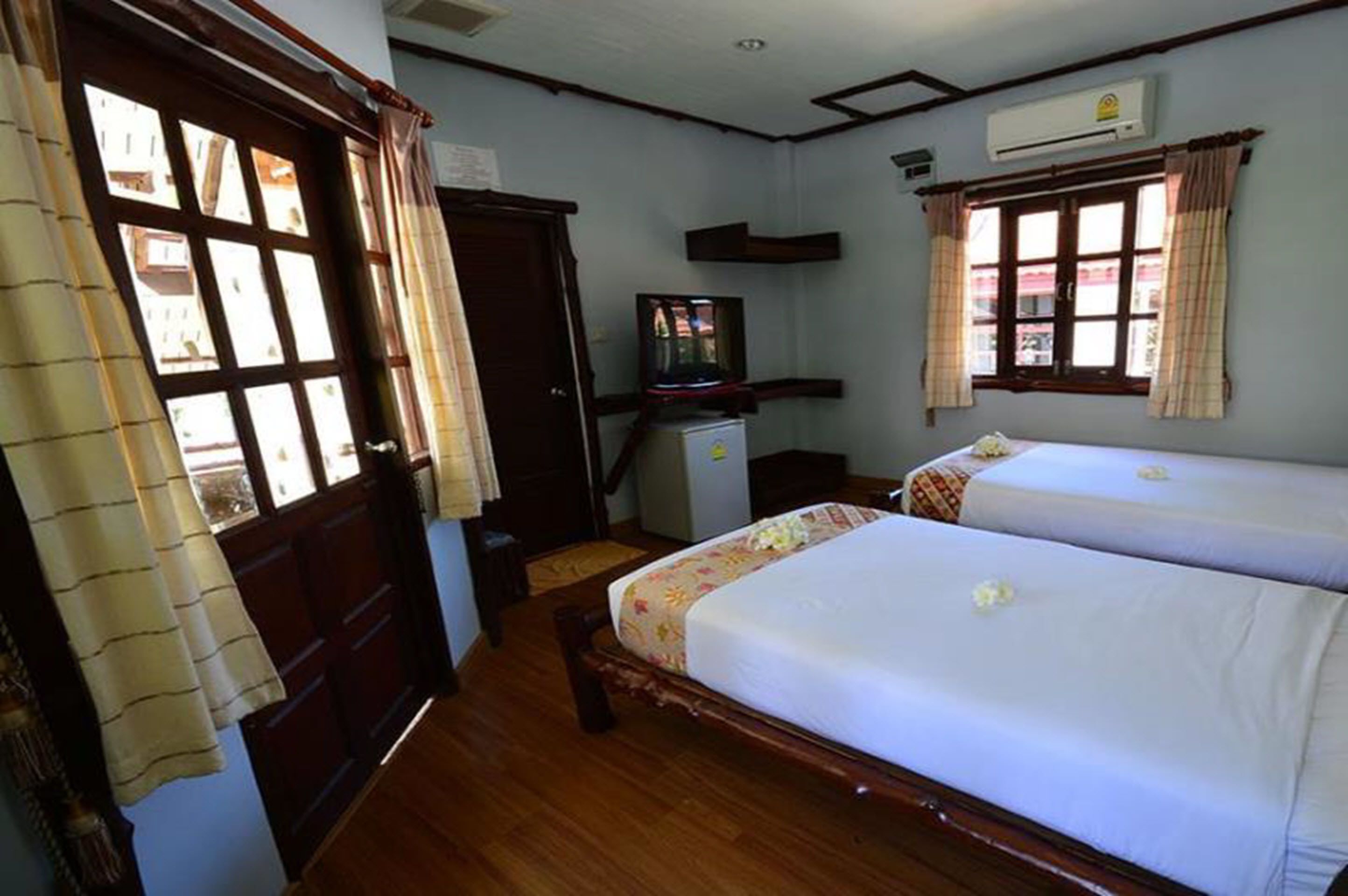 A room with two single beds, a wooden wardrobe, a small desk with a television, and a window with curtains.