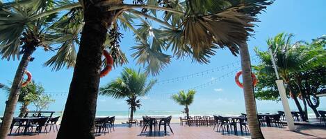 Un patio frente a la playa con mesas y sillas, palmeras y vista al océano.