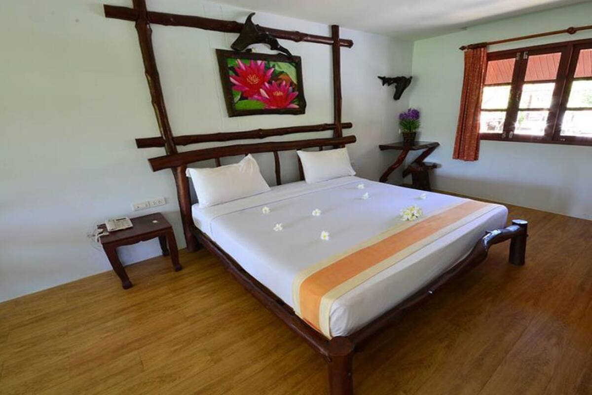 A wooden bed with white bedding and a decorative orange stripe, a small bedside table with a phone, a wall-mounted picture of a flower, and a window with curtains.
