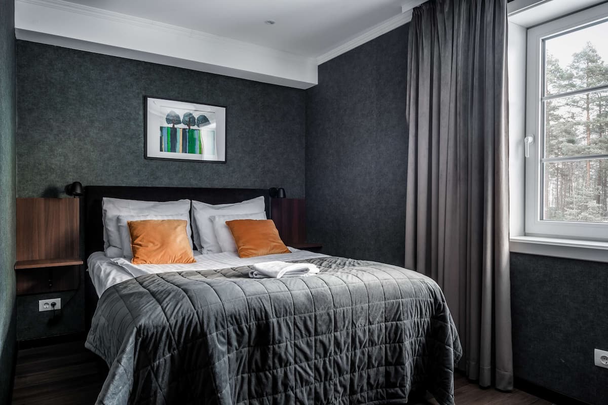 Una cama bien hecha con colcha gris, dos almohadas y una manta blanca. Encima de la cama hay una obra de arte enmarcada, y se ve una ventana con cortinas a la derecha.