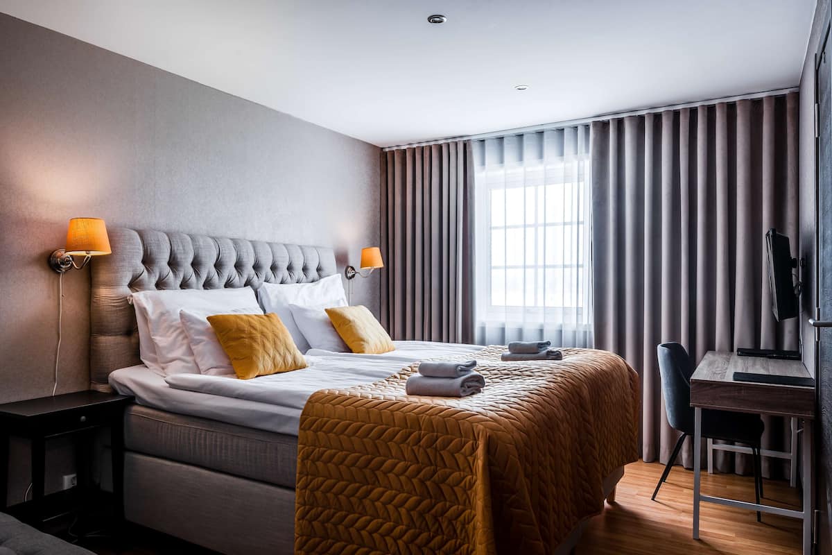 Una habitación de hotel con cabecera capitonada, cama con ropa de cama blanca y mostaza, un escritorio con silla y un ventanal con cortinas.