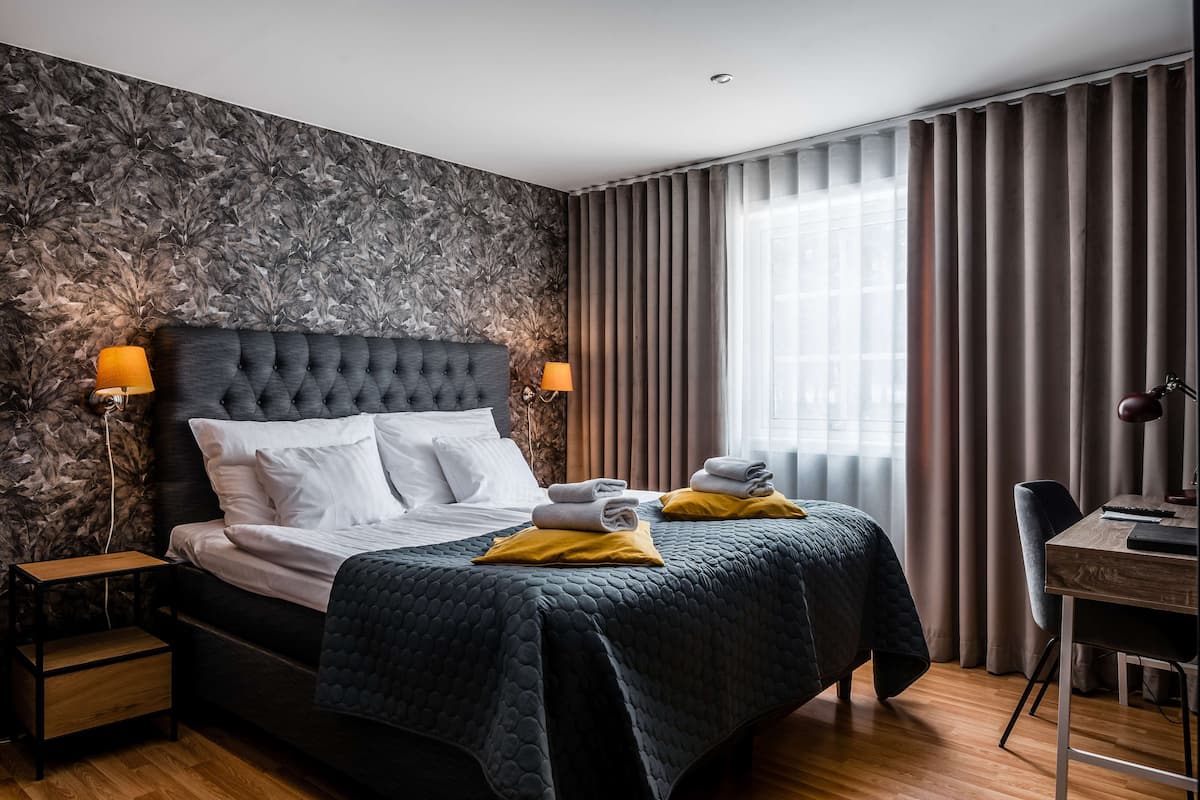 Una habitación de hotel moderna con una cama grande, dos lámparas de noche, un escritorio de madera y vistas de la ciudad a través de amplias ventanas.