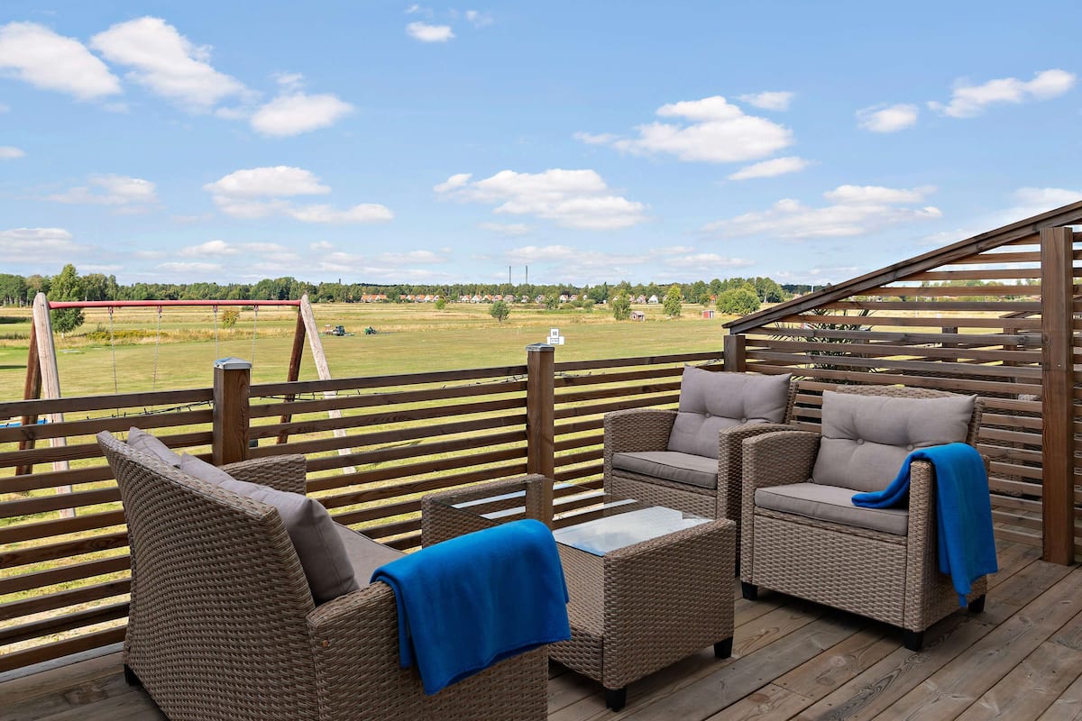 Una terraza de madera con muebles de mimbre y vistas a un paisaje rural.