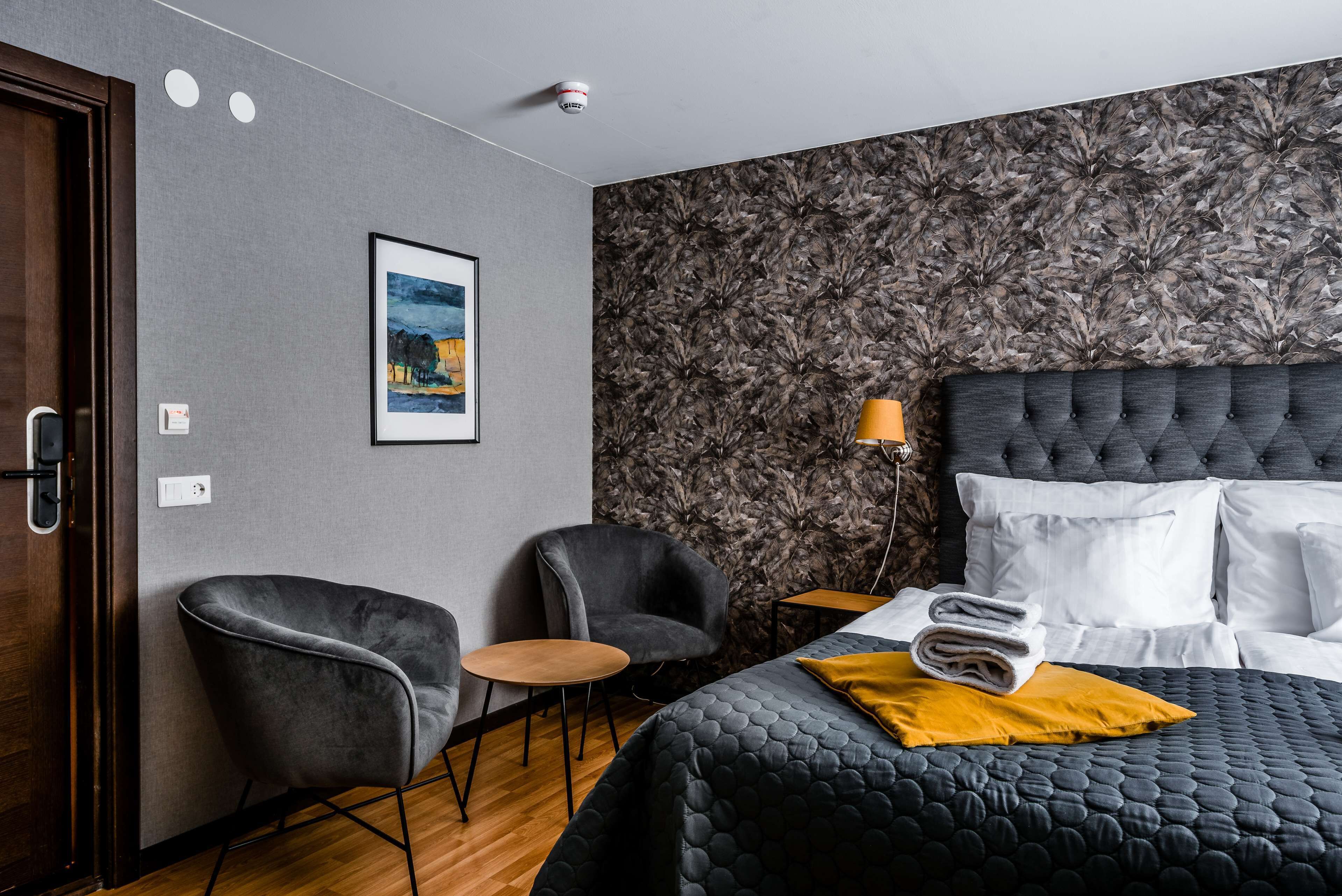 Habitación de hotel con cama, dos sillones, una mesita, un cuadro en la pared y un papel tapiz estampado.