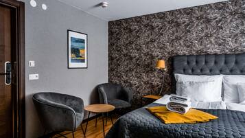 Quarto de hotel com uma cama, duas poltronas, uma mesa pequena, um quadro na parede e papel de parede estampado.