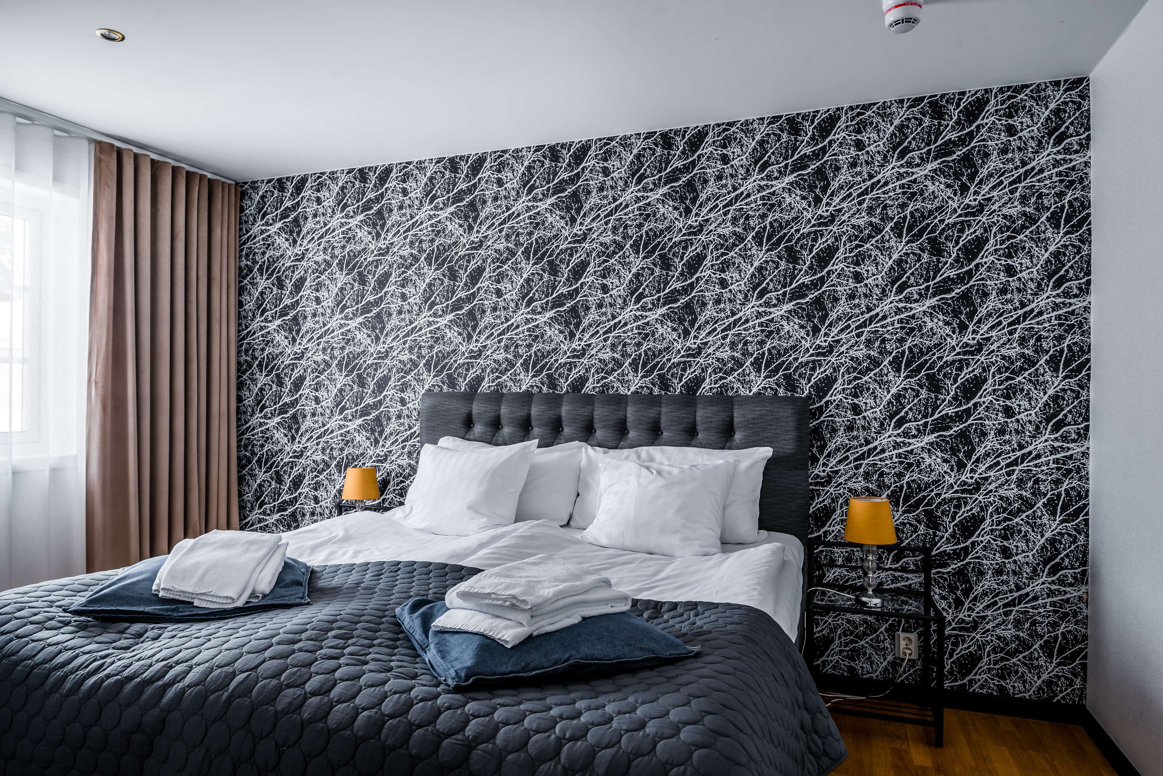 En snyggt bäddad säng med vita sängkläder, ett mörkt, vadderat sängöverkast och två kuddar. Vid sängen står ett litet bord med en lampa och på väggen sitter en hylla. Rummet har en modern design med ett huvudgavels med knäppning och en mönstrad tapet.