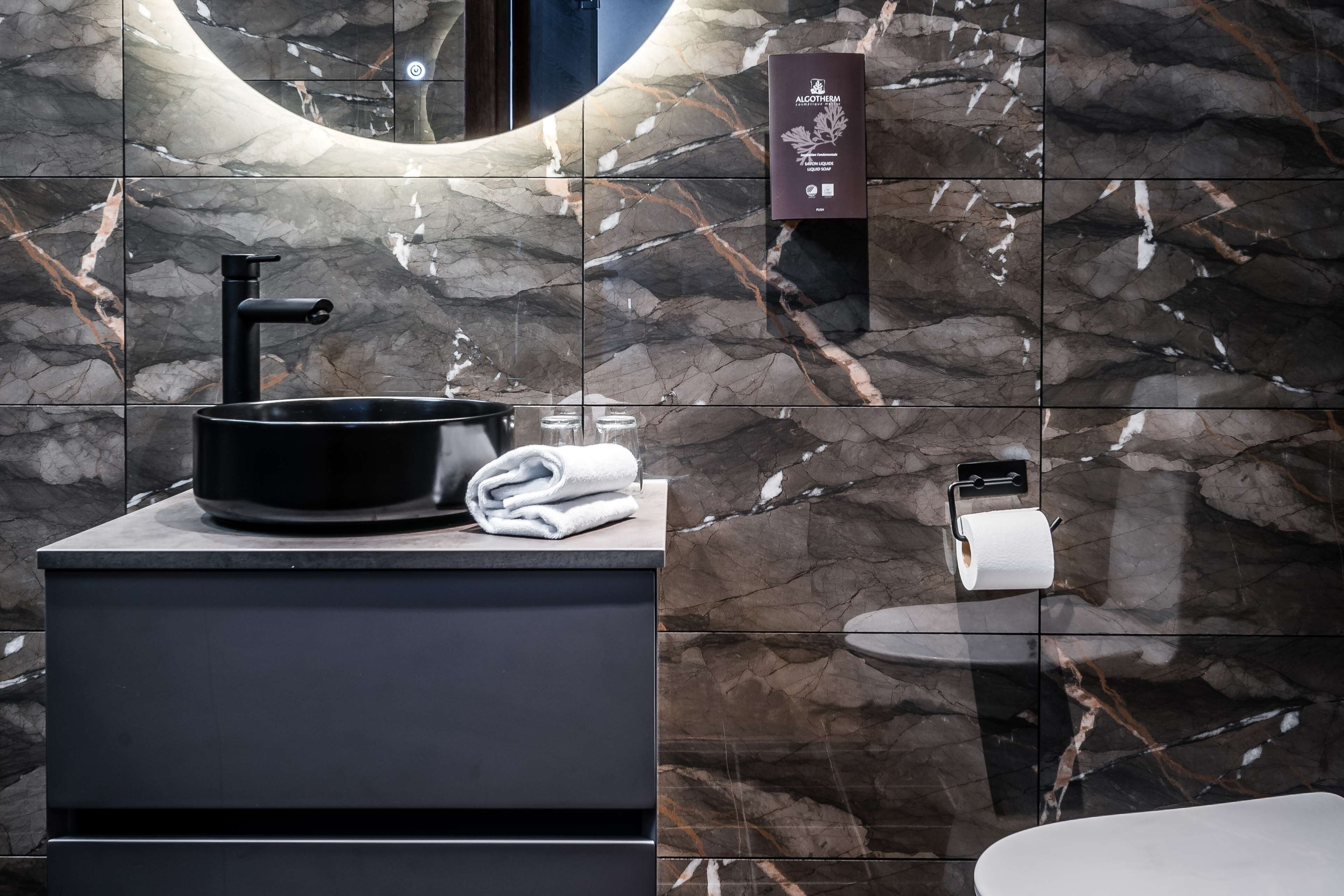 A modern bathroom with a black sink, marble tiles, a towel, and a toilet paper holder.