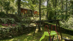 A wooden cabin with a thatched roof, surrounded by lush greenery and a stone wall.