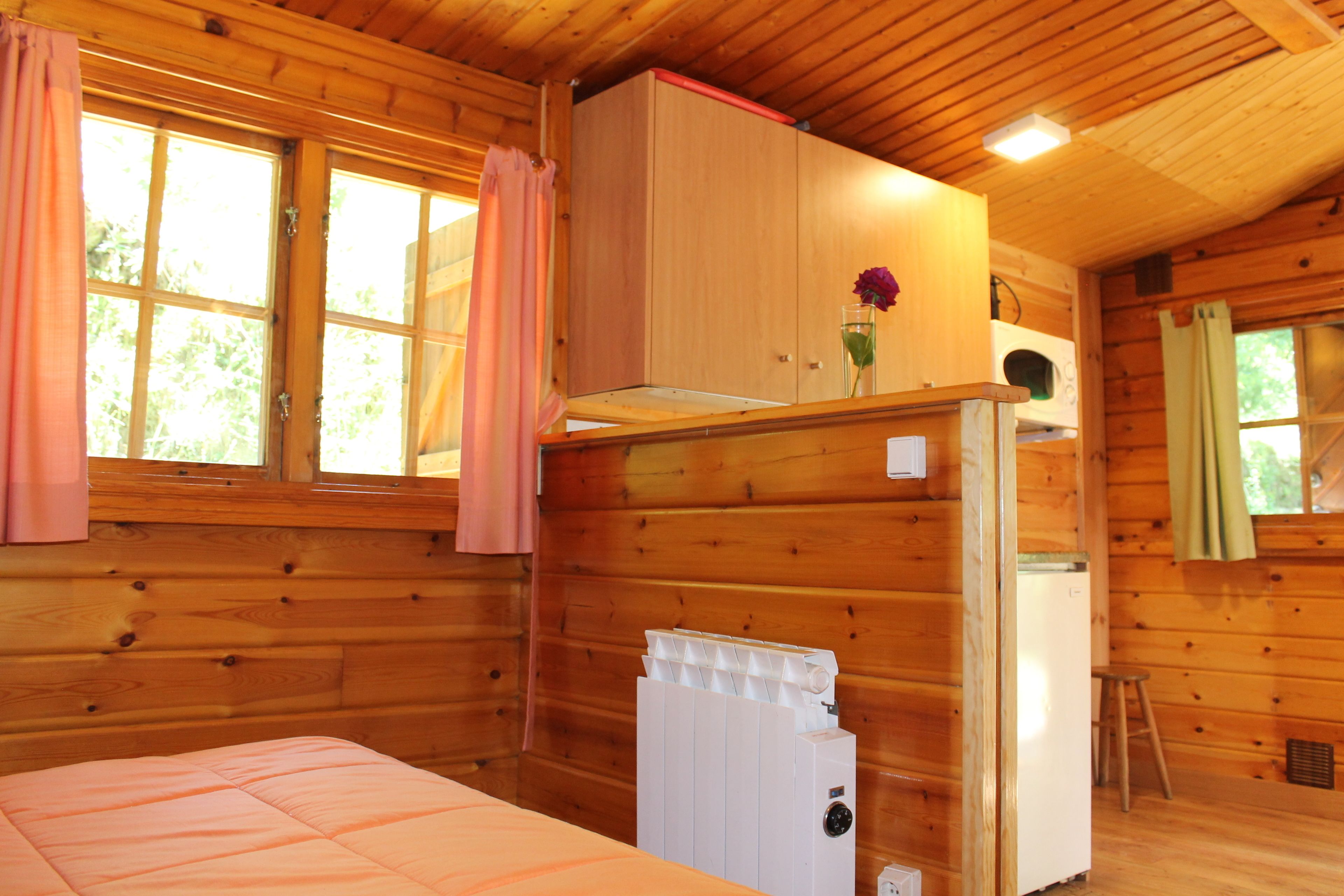 Ein Zimmer in einer Holzhütte mit einem Bett, einem weißen Heizkörper, einem kleinen Kühlschrank und einem Fenster mit Vorhängen.