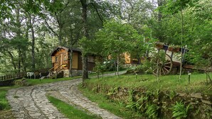 A stone pathway leading to a rustic wooden cabin with a wooden cart and a small bridge over a stream.