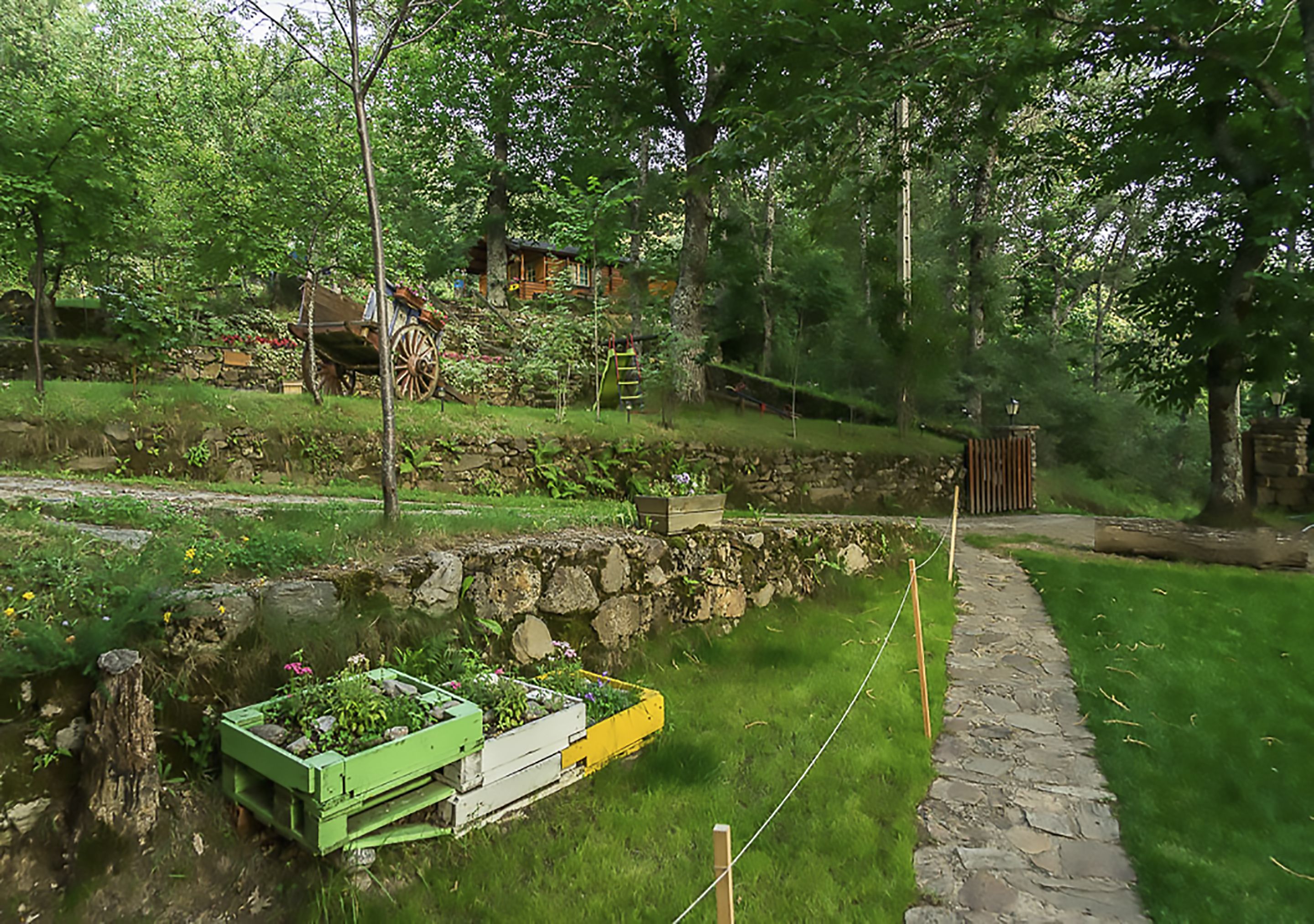 Una cabaña rústica rodeada de abundante vegetación, un muro de piedra y un pequeño arroyo.