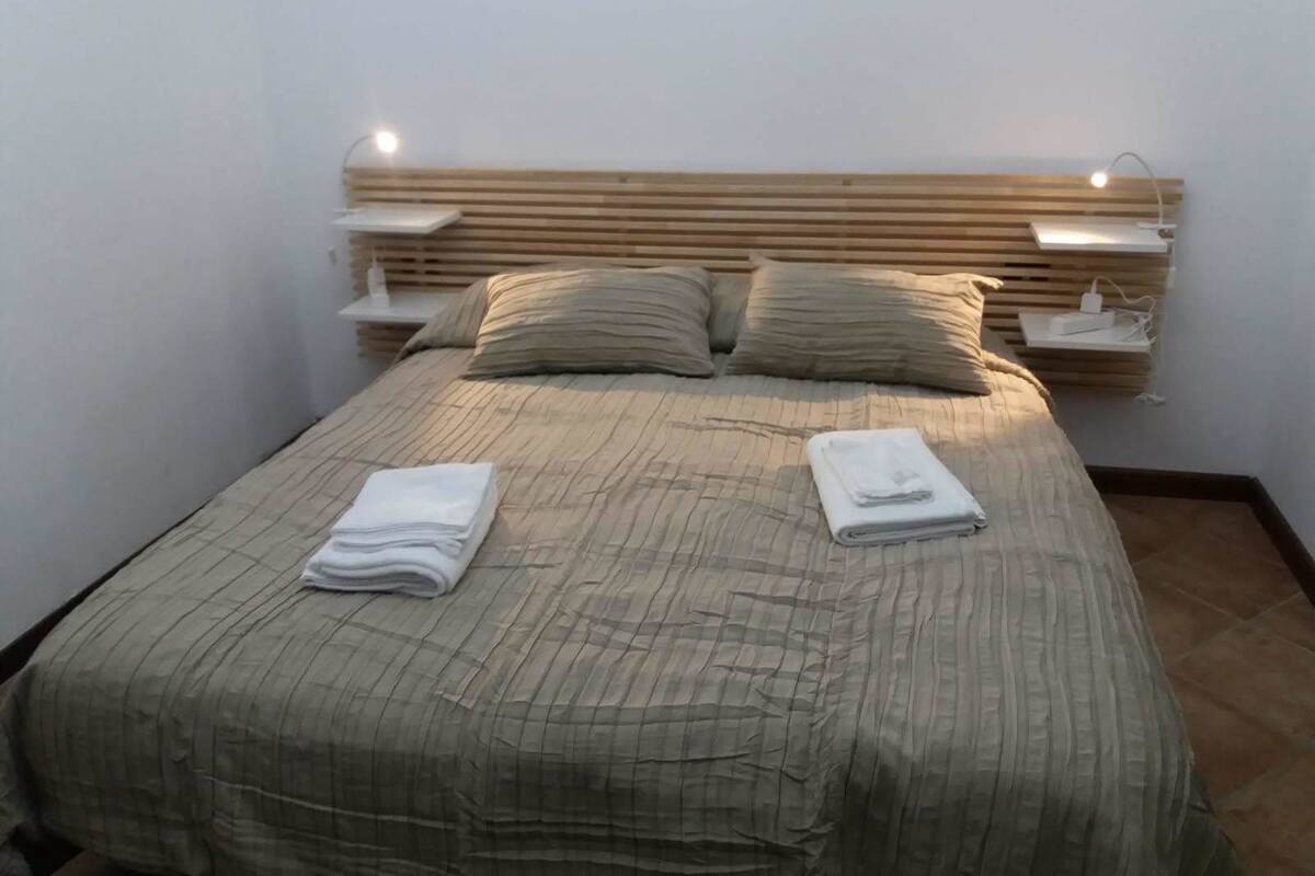 A neatly made bed with a striped bedspread, two pillows, and folded towels on the bed. There are bedside tables with lamps and a wall-mounted shelf above the bed.