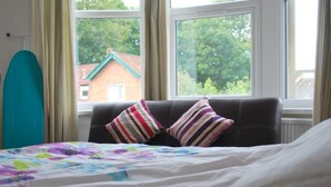 A bedroom with a bed, striped pillows, a window with a view of a house, and a teal surfboard leaning against the wall. - Clift Guest House (Bristol)