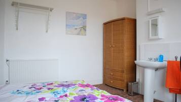 A bedroom with a bed, a wardrobe, a radiator, a painting, a sink, and a clock.