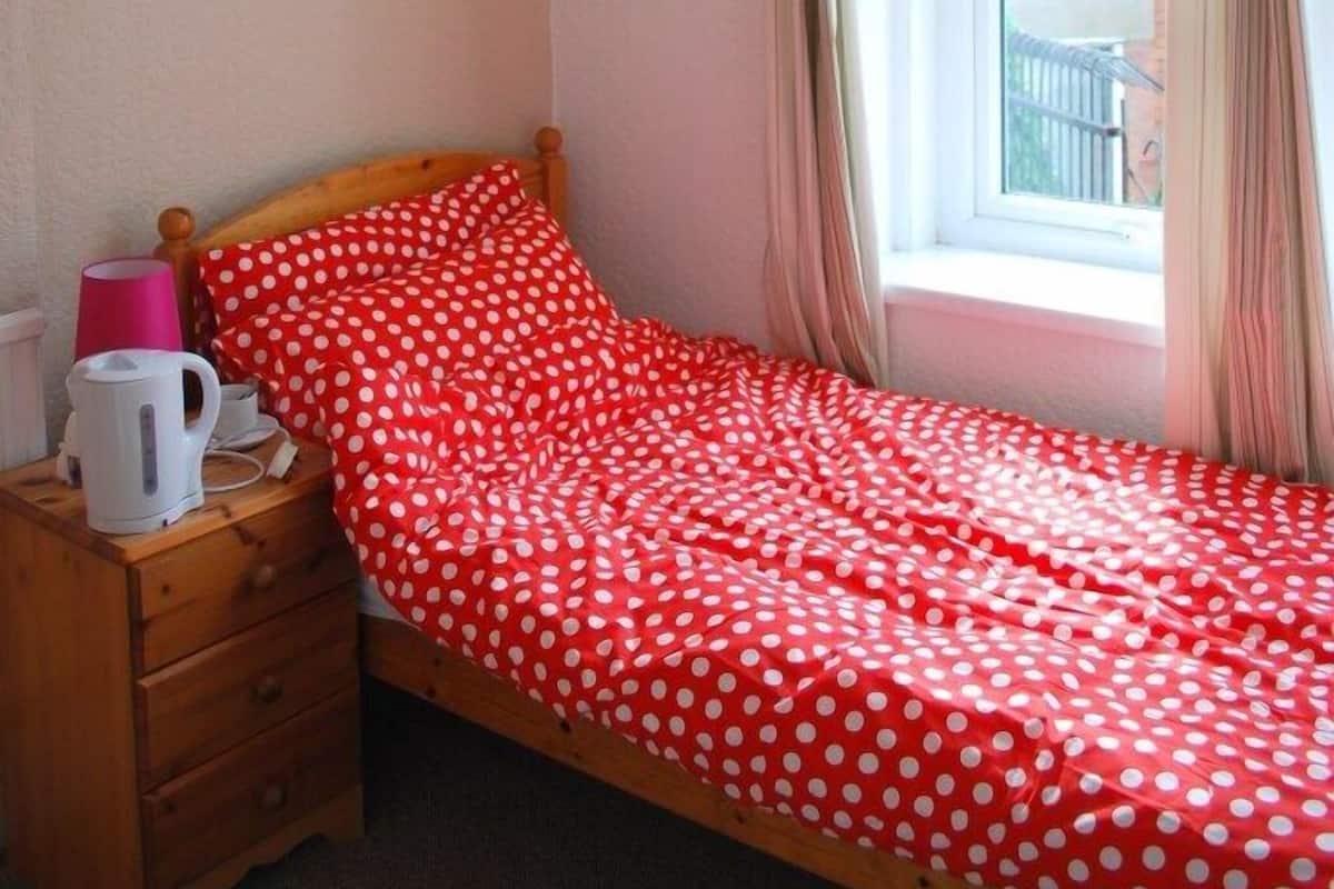 A single bed with a red polka dot bedspread, a wooden nightstand with a pink cup and a white kettle, a painting on the wall, and a window with curtains.