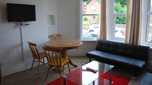 A living room with a round wooden table, two wooden chairs, a black leather sofa, a glass coffee table, and a television mounted on the wall. - Clift Guest House (Bristol)