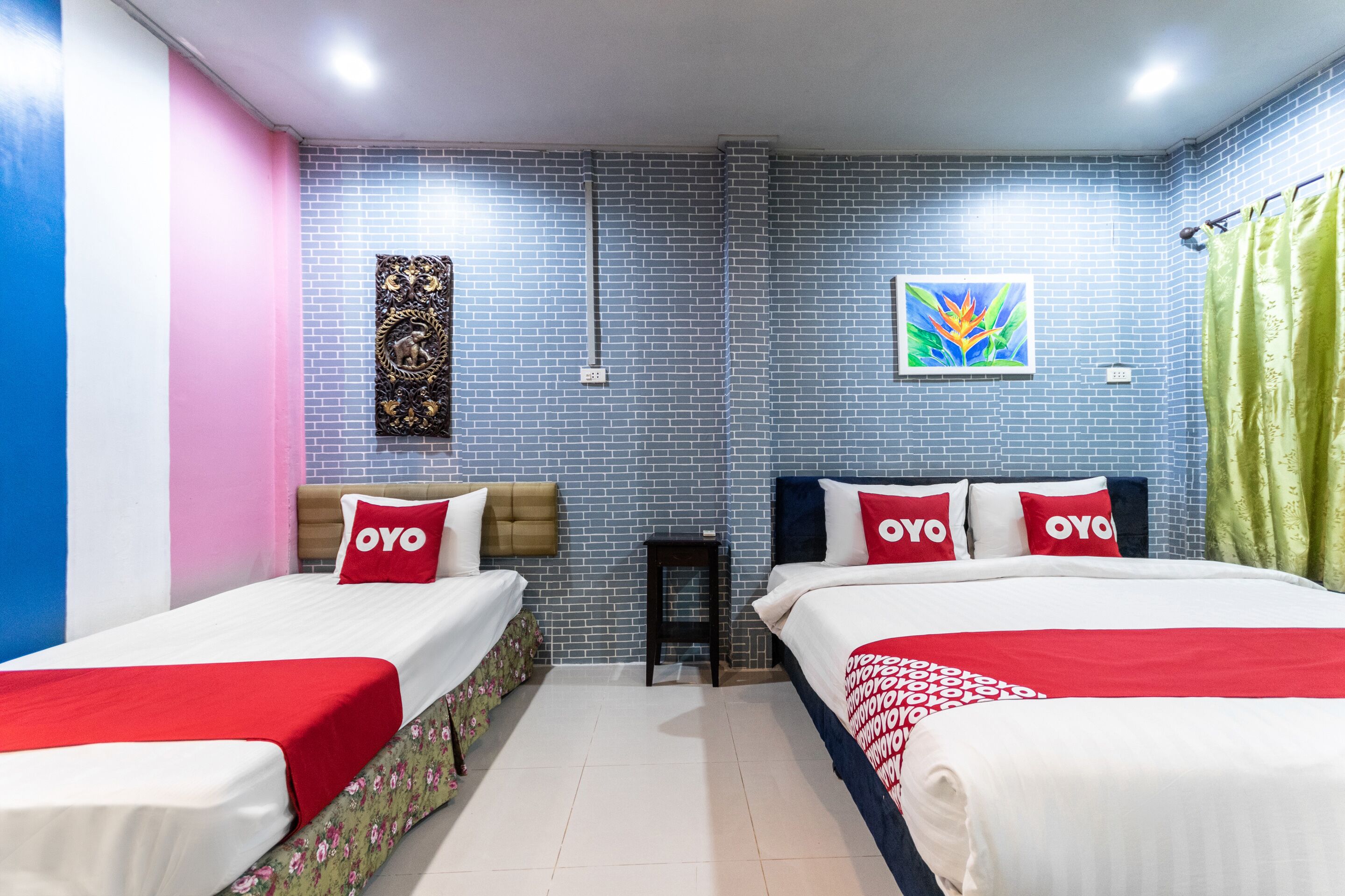 Een hotelkamer met twee bedden, elk met een rode en witte spreidek en kussens met de tekst 'OYO'. De kamer heeft een blauwe bakstenen muur, een kleine tafel en een fotolijst.
