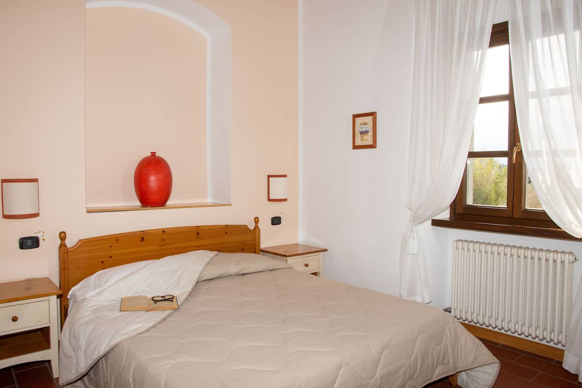 Une chambre à coucher avec un lit en bois, une table de chevet, un vase rouge, une fenêtre avec des rideaux et un radiateur.