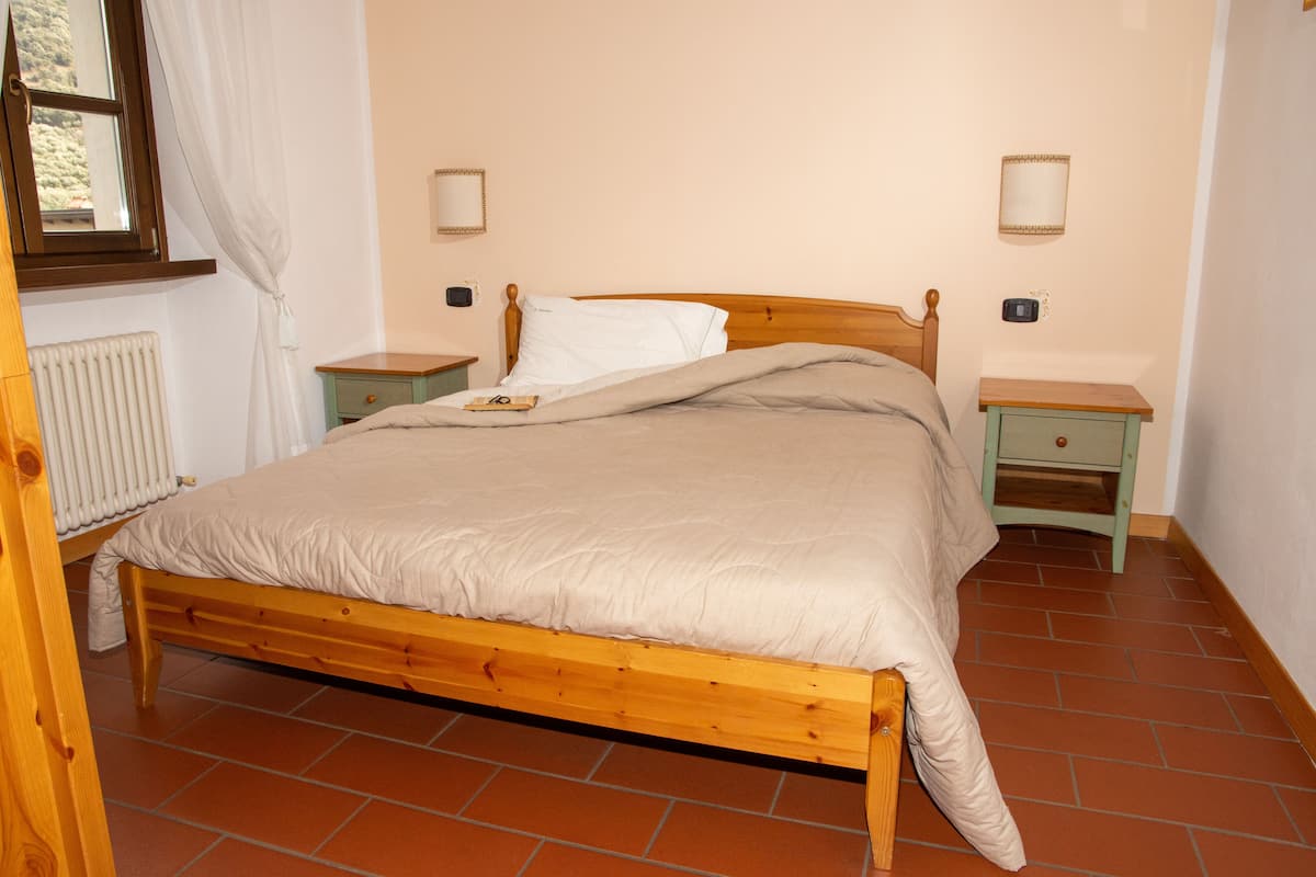 Un lit en bois recouvert d’une couverture beige, deux tables de chevet, une commode, un radiateur et une fenêtre avec des rideaux.
