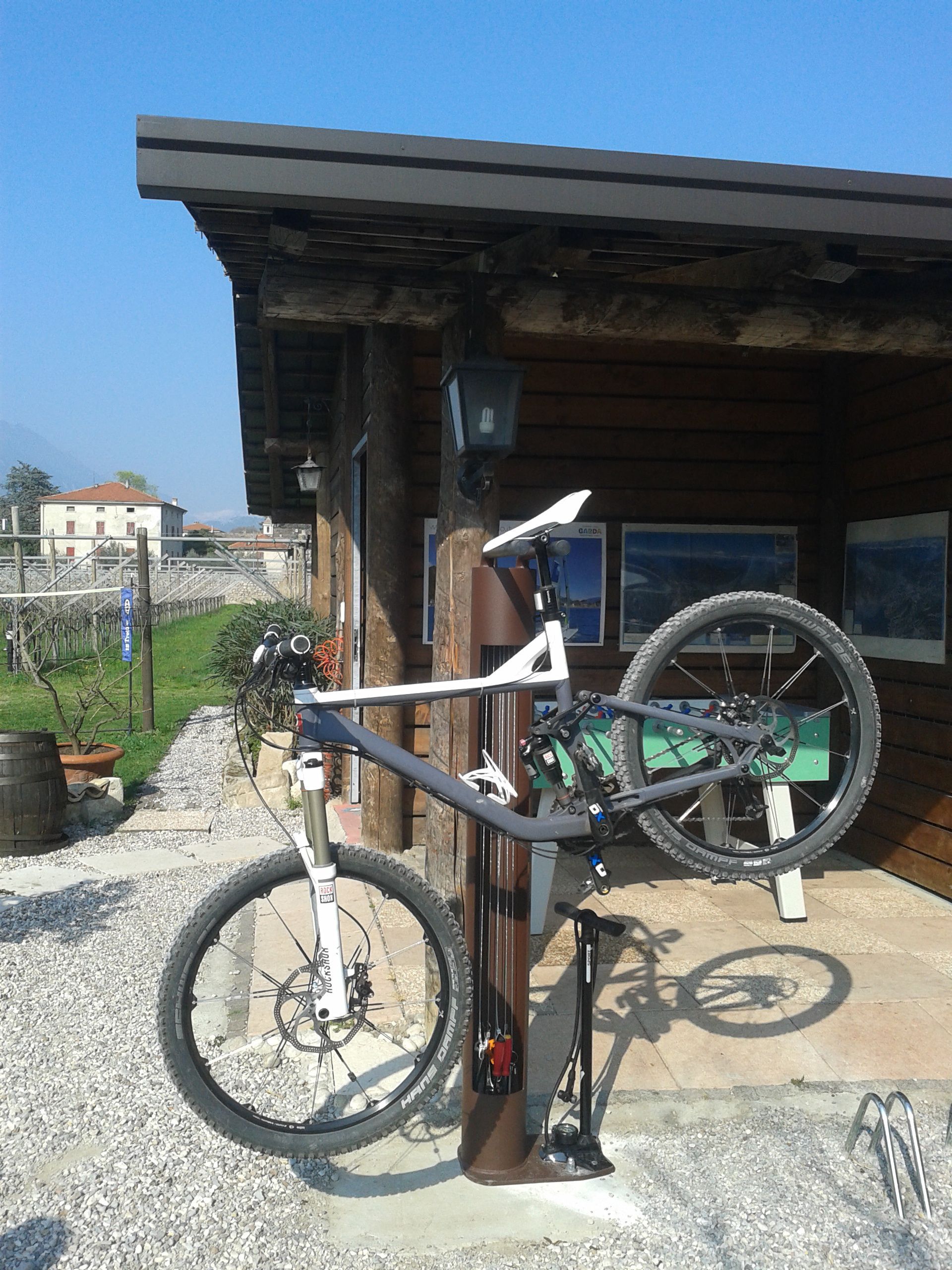 Un vélo tout-terrain est garé contre un bâtiment en bois, avec un chemin de gravier et une clôture en bois en arrière-plan.