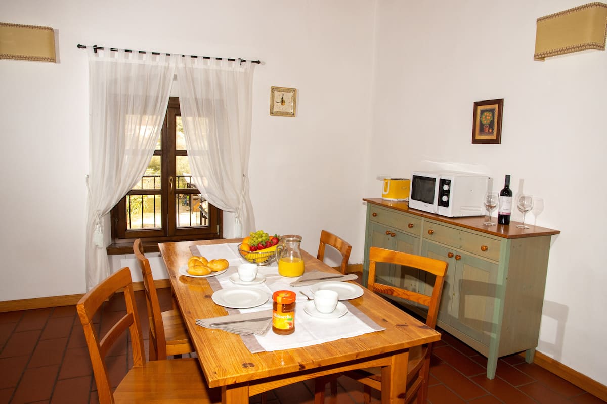 Un coin repas avec une table en bois, des chaises, un panier de fruits, une carafe de jus d’orange et un meuble contenant un four à micro-ondes.