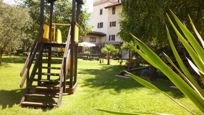 Un terrain de jeux avec un toboggan en bois et une structure d’escalade, entouré de pelouse et d’arbres, avec un bâtiment visible au fond.