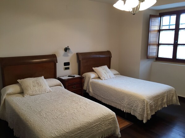 A hotel room with two beds, a nightstand, a wall-mounted lamp, and a window with blinds.