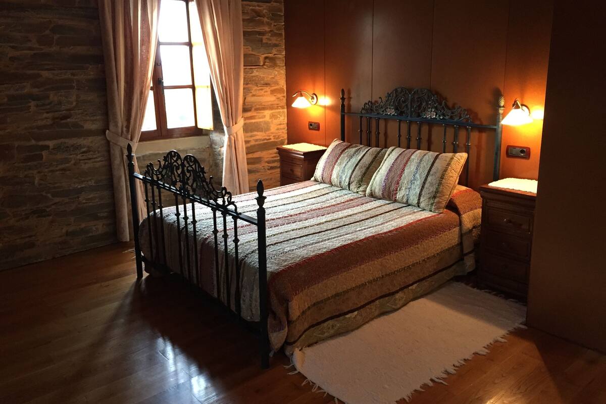 Un dormitorio con pared de piedra, estructura de cama de madera, colcha a rayas y dos lámparas de noche.
