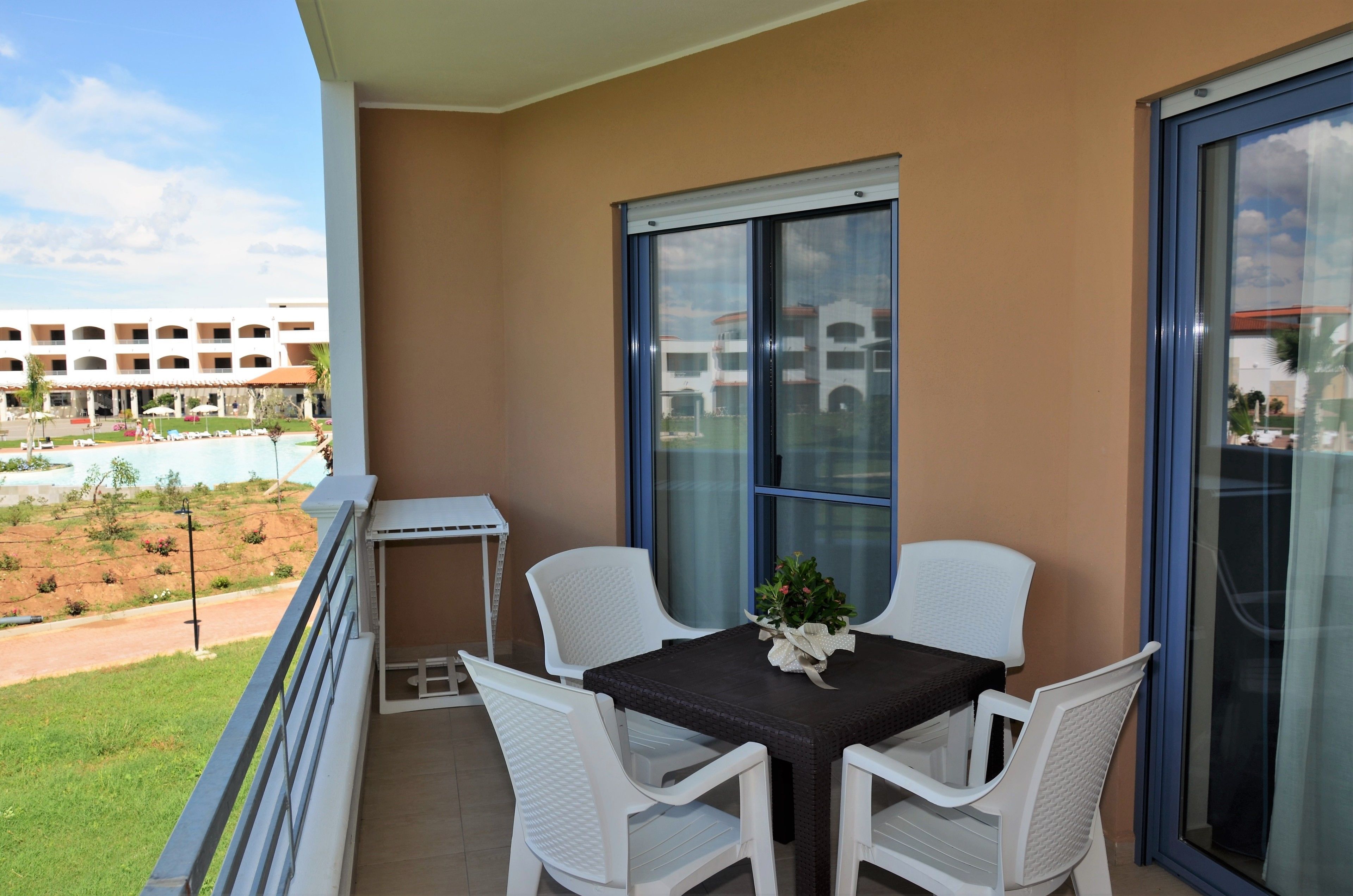 Un balcone con tavolo e sedie, una finestra che si affaccia su un edificio e una piscina, e un piccolo tavolino con una pianta.