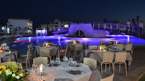 Un área de comedor al aire libre con mesas dispuestas para los huéspedes, con vista a una masa de agua con un puente y fuentes iluminadas.