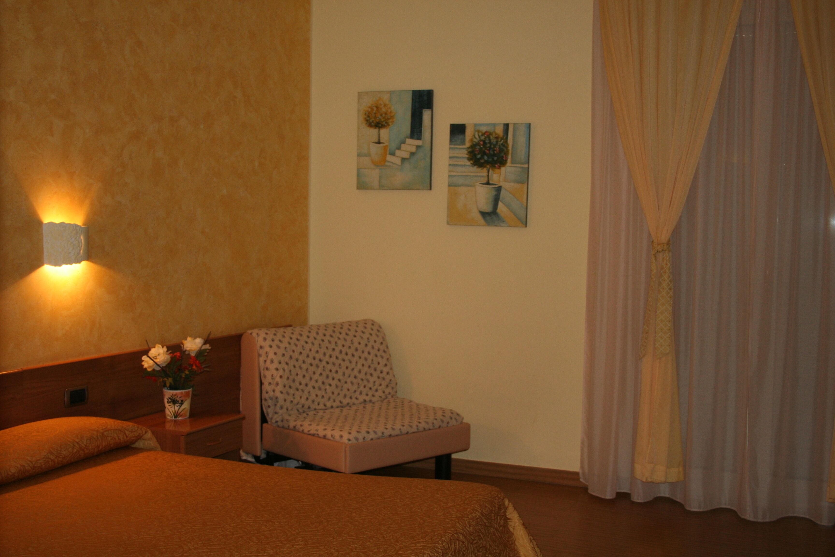 Een hotelkamer met een bed, een stoel, een nachtkastje met een bloevas en twee schilderijen aan de muur.