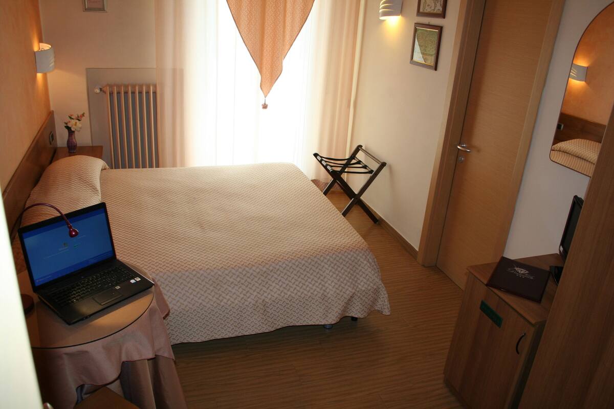 Habitación de hotel con cama, escritorio, laptop, radiador, silla y armario.