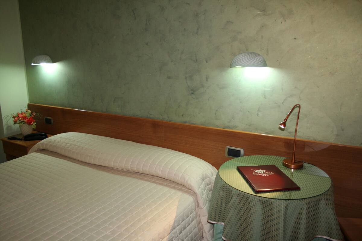 Habitación de hotel con cama, mesita de noche con lámpara, una pequeña mesa redonda con un libro y dos lámparas de pared.
