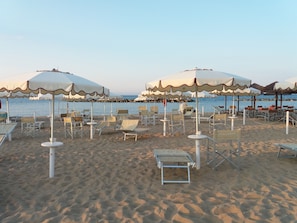 Een strand met witte parasols en ligstoelen, een bord met de tekst 'Welkom op het strand' en de oceaan op de achtergrond.