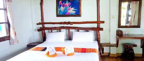A wooden-framed bed with white bedding, a painting of flowers above it, and a wooden desk with a mirror to the right.
