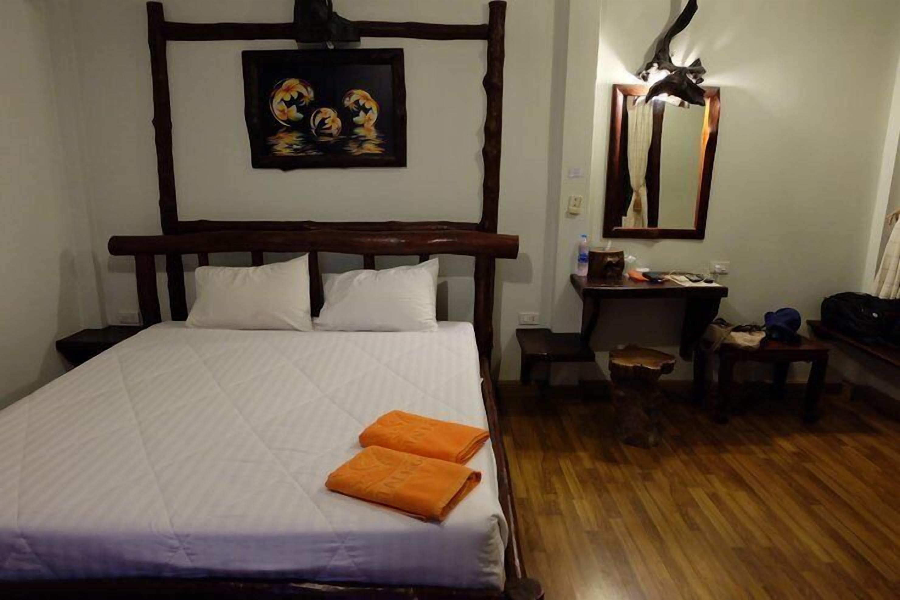 A bedroom with a wooden bed frame, white bedding, an orange towel, a painting above the bed, a mirror, and a desk with a chair.