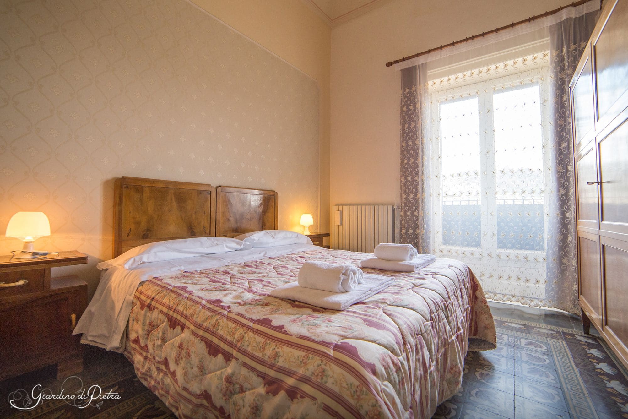 A hotel room with a large bed, a wooden headboard, a nightstand with a lamp, and a window with curtains.