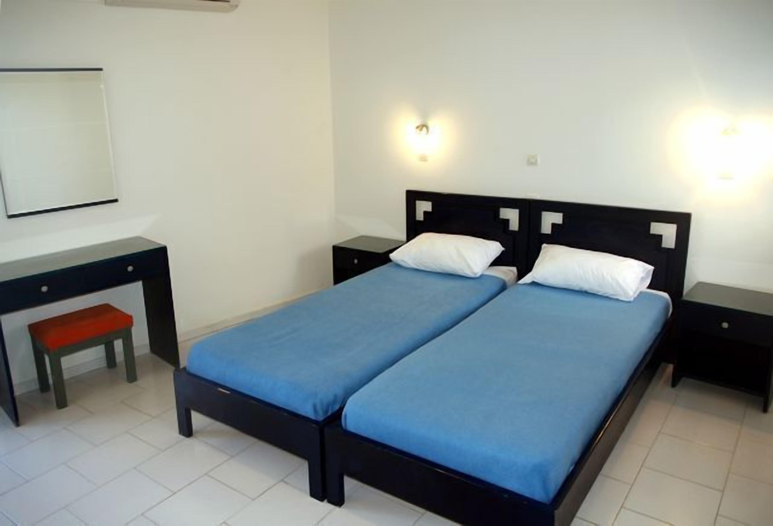 Uma cama de casal com colcha azul, duas mesas de cabeceira, um pequeno banco vermelho e um espelho branco fixado na parede.