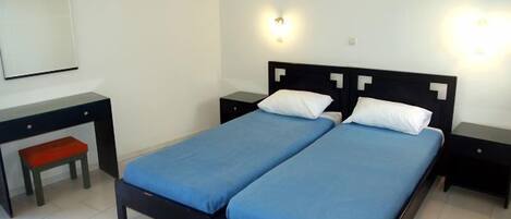 Uma cama de casal com colcha azul, duas mesas de cabeceira, um pequeno banco vermelho e um espelho branco fixado na parede.