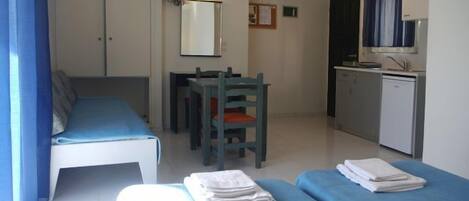 A small, well-lit room with a bed, a desk, a chair, and a kitchenette.