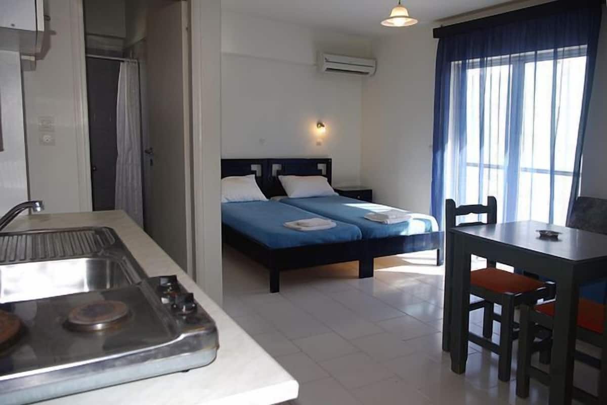 A compact hotel room with a kitchenette, a bed with two pillows, a dining table with chairs, and a large window with curtains.