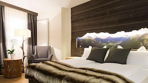 A hotel room with a large bed, a chair, a wooden side table, a lamp, and a mountain landscape painting on the wall.