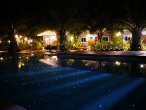 A nighttime view of a pool with reflections of lights, surrounded by well-lit buildings with a bench and plants.