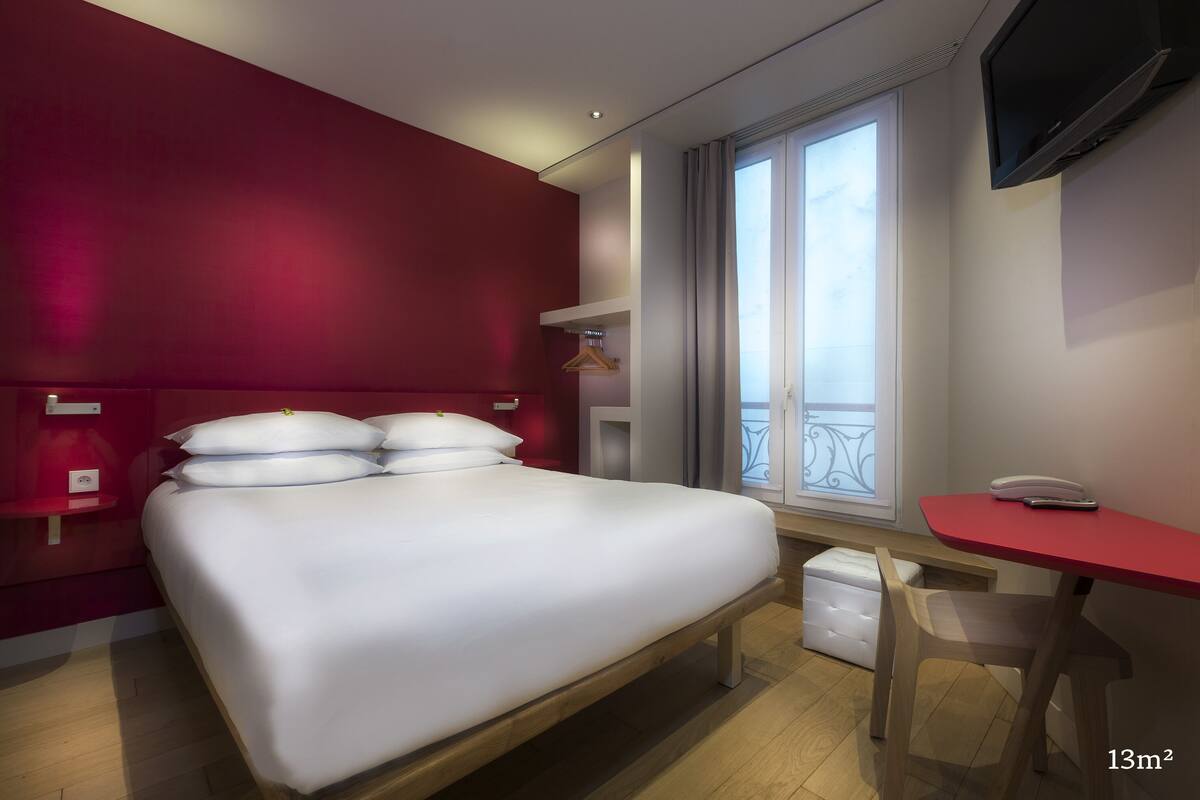 A hotel room with a bed, a desk, a chair, a TV, and a window with a balcony view.