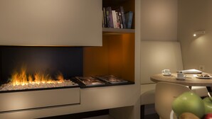 A modern room with a built-in fireplace, a bookshelf, a dining table with a cup and muffin, and a fruit bowl. - Hotel Andrea (Paris)