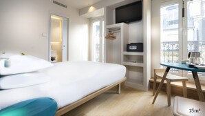 A modern hotel room with a bed, a desk, and a chair. There is a TV mounted on the wall, and a window with a view of the outside. - Hotel Andrea (Paris)