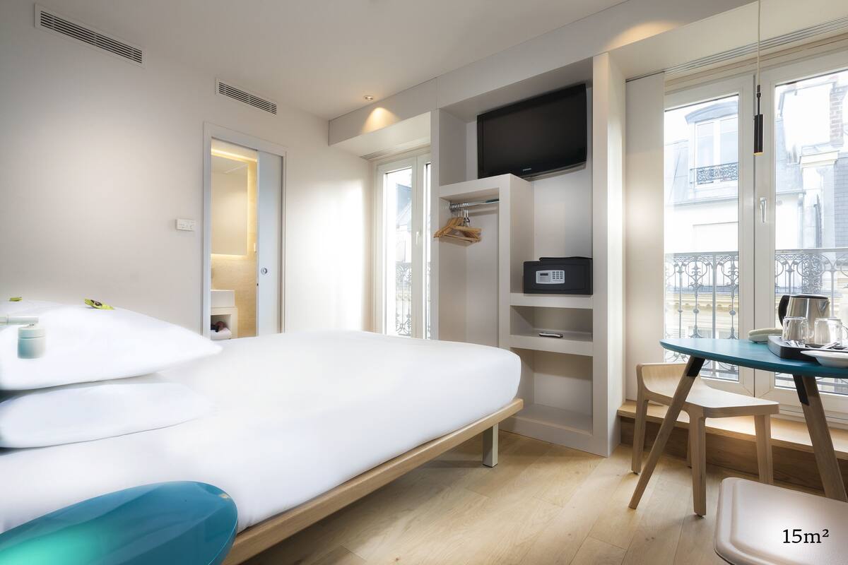 A modern hotel room with a bed, a desk, and a chair. There is a TV mounted on the wall, and a window with a view of the outside.