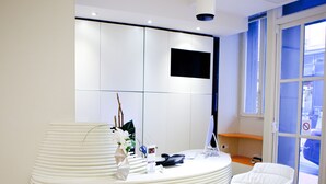 A reception area with a modern, curved reception desk, a white chair, and a flat-screen TV mounted on the wall. - Hotel Andrea (Paris)