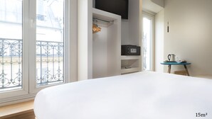 A hotel room with a large bed, a small table with a kettle, a shelf with a TV and a remote control, and a balcony with a railing. - Hotel Andrea (Paris)